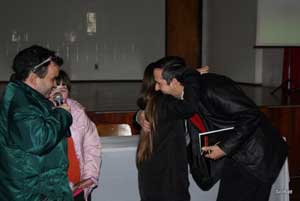 Nesta foto, Alex Garcia segura o microfone, sorri. Na frente dele o palestrante Marcos Henkes abraça uma jovem de cabelos castanhos, lisos e longos. Ele segura um livro na mão esquerda. Ao lado esquerdo de Alex, uma jovem da ONG DOWN observa. Todos estão usando roupas próprias de frio.
