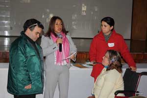 Foto com quatro pessoas. De pé Alex Garcia, Dra. Scheila Lima, apoiadora Aline e Fátima Braum, da Associação dos Amigos Deficientes, sentada em cadeira de Rodas. Dra Scheila fala ao microfone e tem um documento nas mãos.